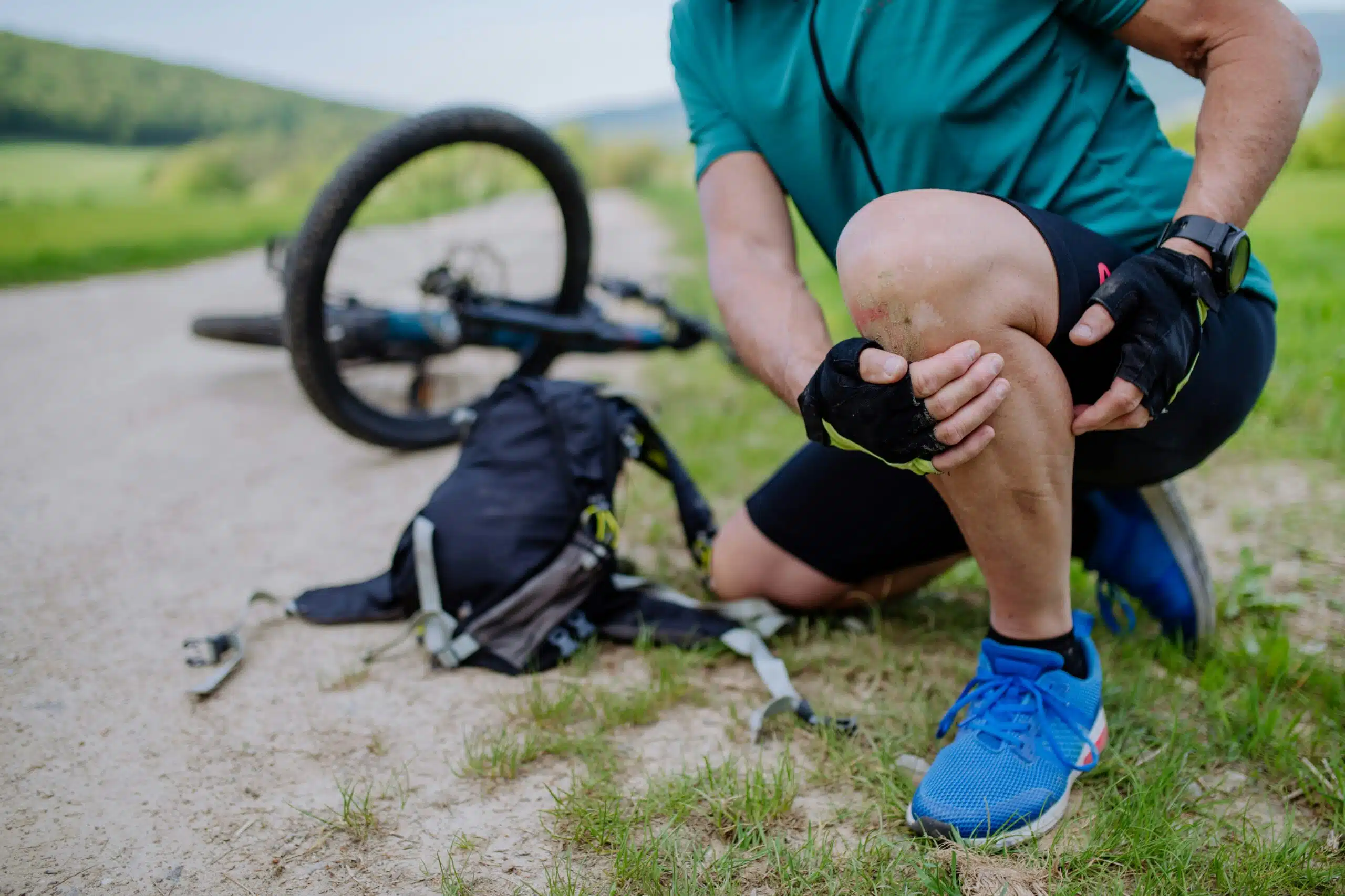 Cyclist kneeling on a dirt path, holding a scraped and bruised knee after a fall. A mountain bike and backpack lie on the ground nearby, suggesting a biking accident during an outdoor ride.
