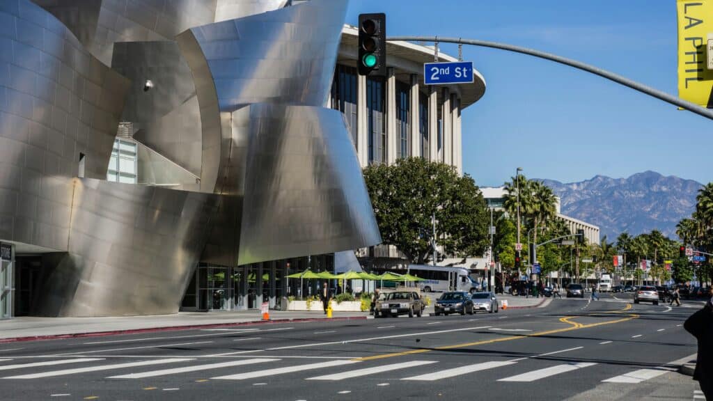 Modern street scene in downtown Los Angeles with the metallic exterior of the Walt Disney Concert Hall on the left, traffic moving along 2nd Street, palm trees lining the road, and mountains visible in the clear background