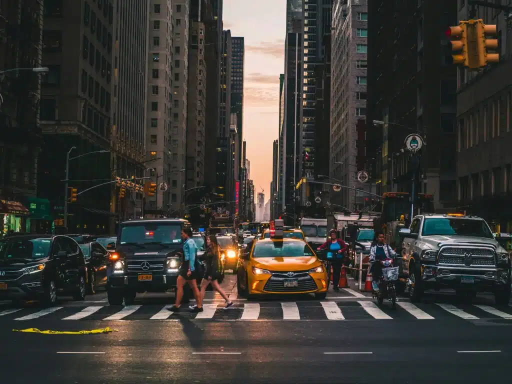 Un concurrido cruce urbano al atardecer, con mucho tráfico y varios peatones cruzando un amplio paso de peatones, rodeado de edificios altos e iluminado por las farolas y los faros de los coches.