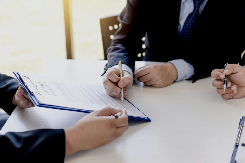 Close-up of three people in business attire sitting at a table, reviewing and signing a contract on a clipboard, symbolizing a formal agreement or legal consultation.