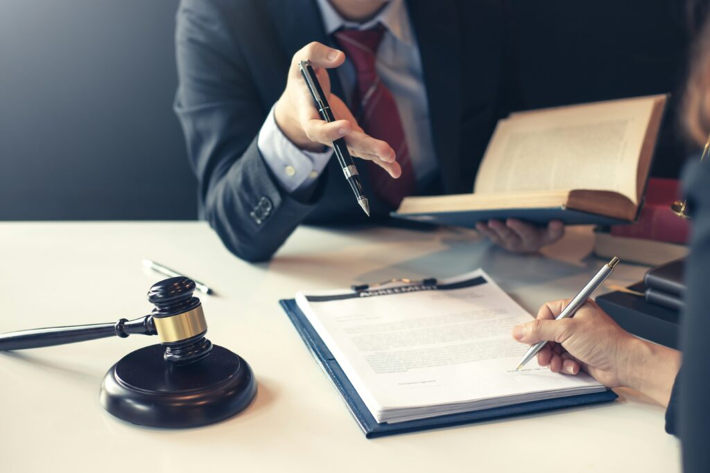 At a desk, two people appear to be going over legal paperwork — one holding a pen and gesturing while referencing an open book, the other signing documents on a clipboard. A judge’s gavel rests nearby, reinforcing the legal or courtroom-re
