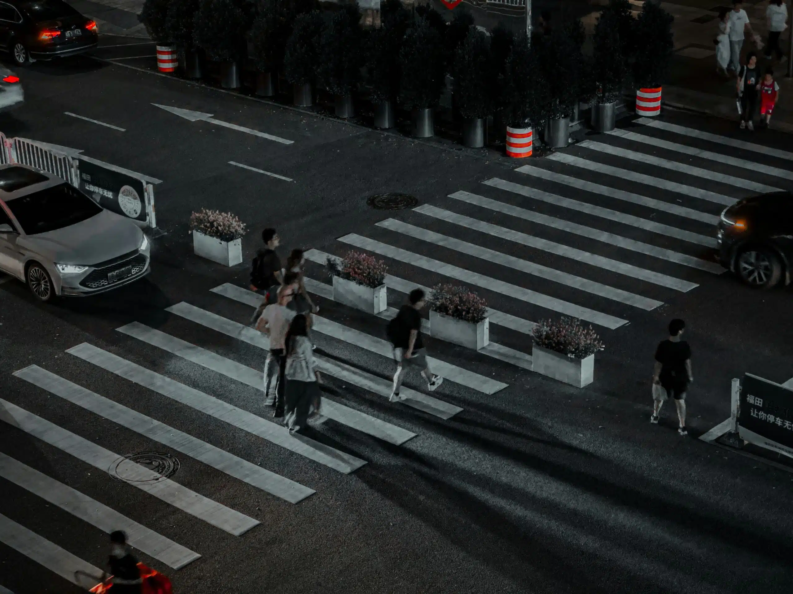 Una escena nocturna en un paso de peatones urbano en la que se ve a varios peatones cruzando unos pasos de peatones anchos y con rayas blancas, iluminados por las farolas y los vehículos que pasan, con jardineras y conos de tráfico colocados entre los carriles.