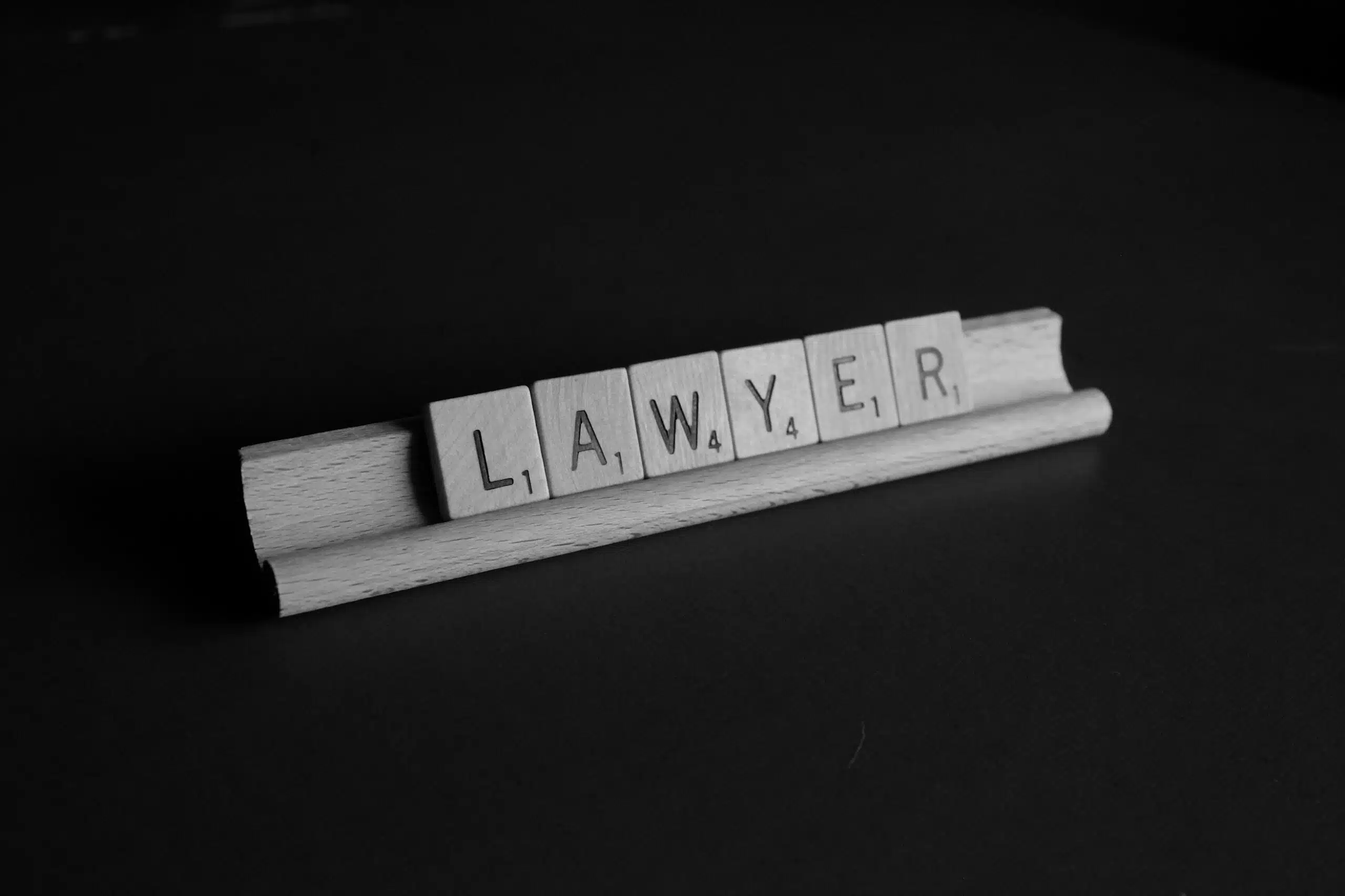 Imagen en blanco y negro de fichas de madera con letras que forman la palabra «LAWYER», dispuestas sobre un pequeño soporte de madera sobre un fondo oscuro, con una iluminación suave que crea un ambiente profesional y minimalista propio del ámbito jurídico.
