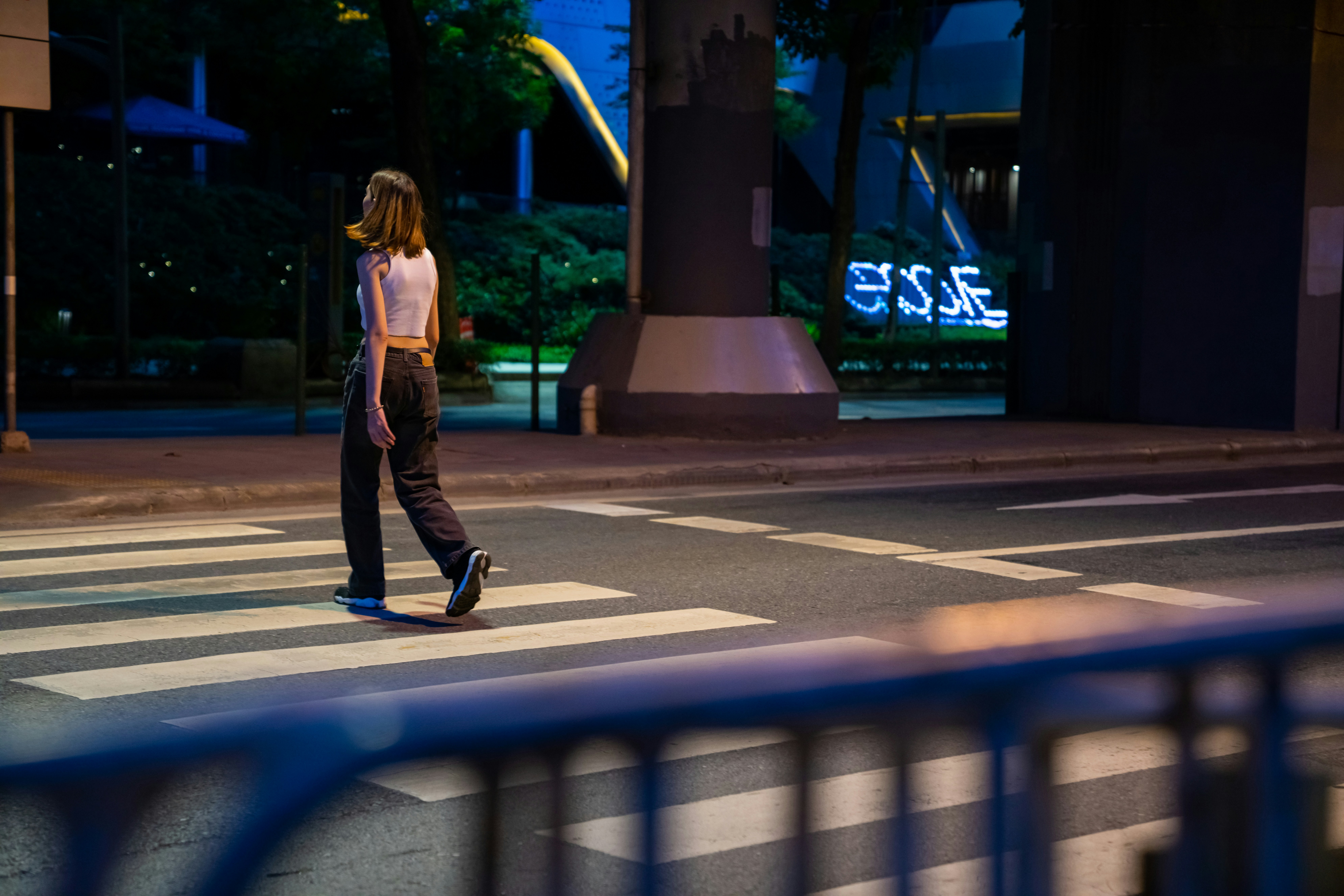 A woman crossing the street at night, illuminated by streetlights, with a cityscape in the background.